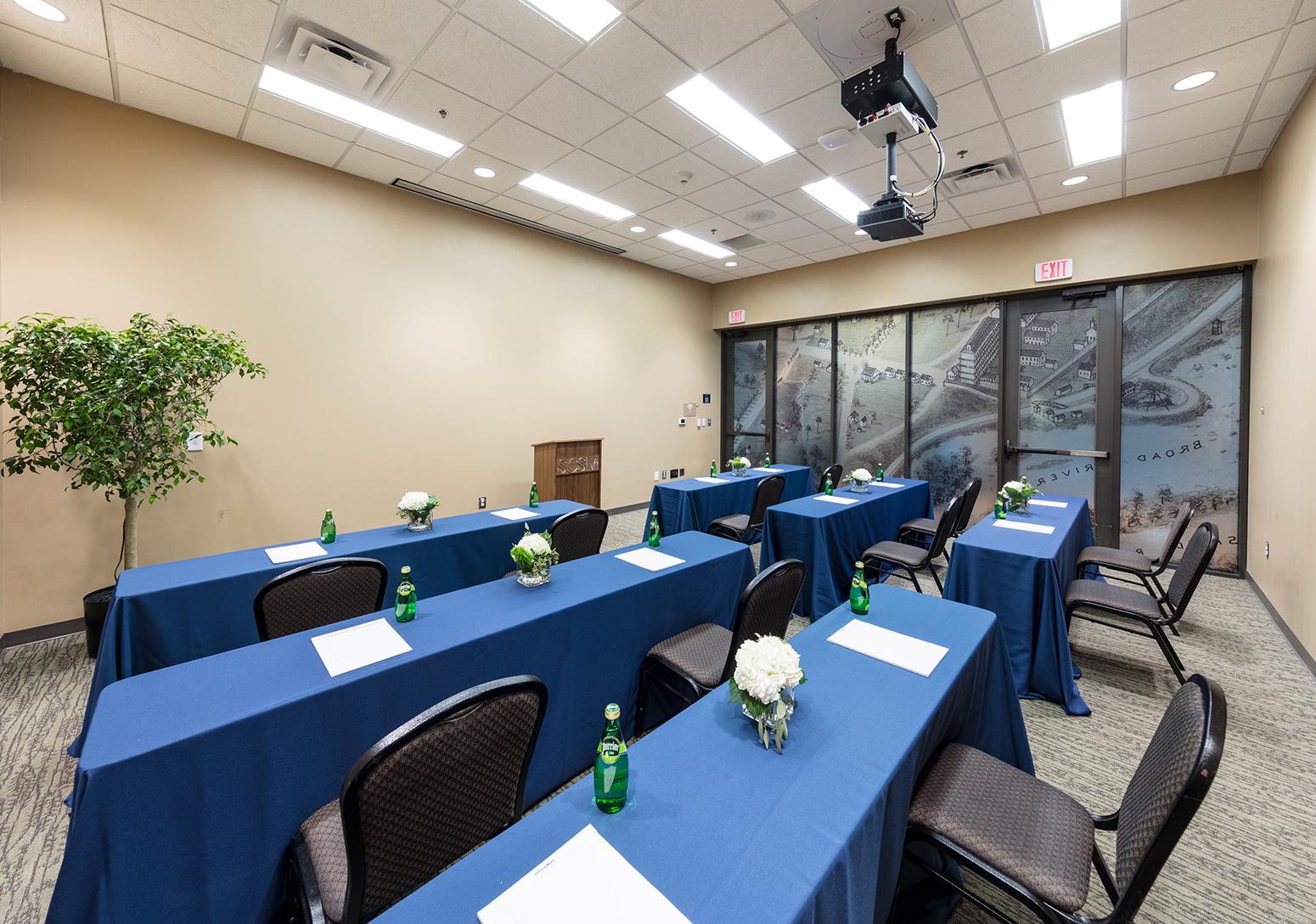 Room with tables with blue table cloths set up facing a speaker's podium