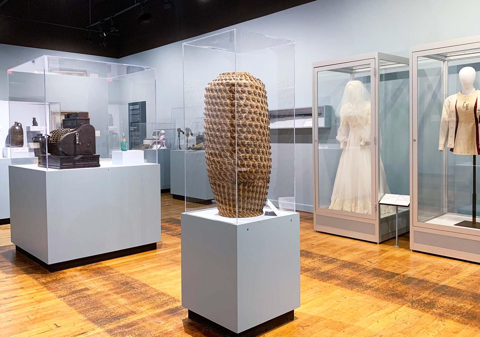Gallery with display cases containing a large, tall narrow vase, pottery, a wedding dress and a jacket