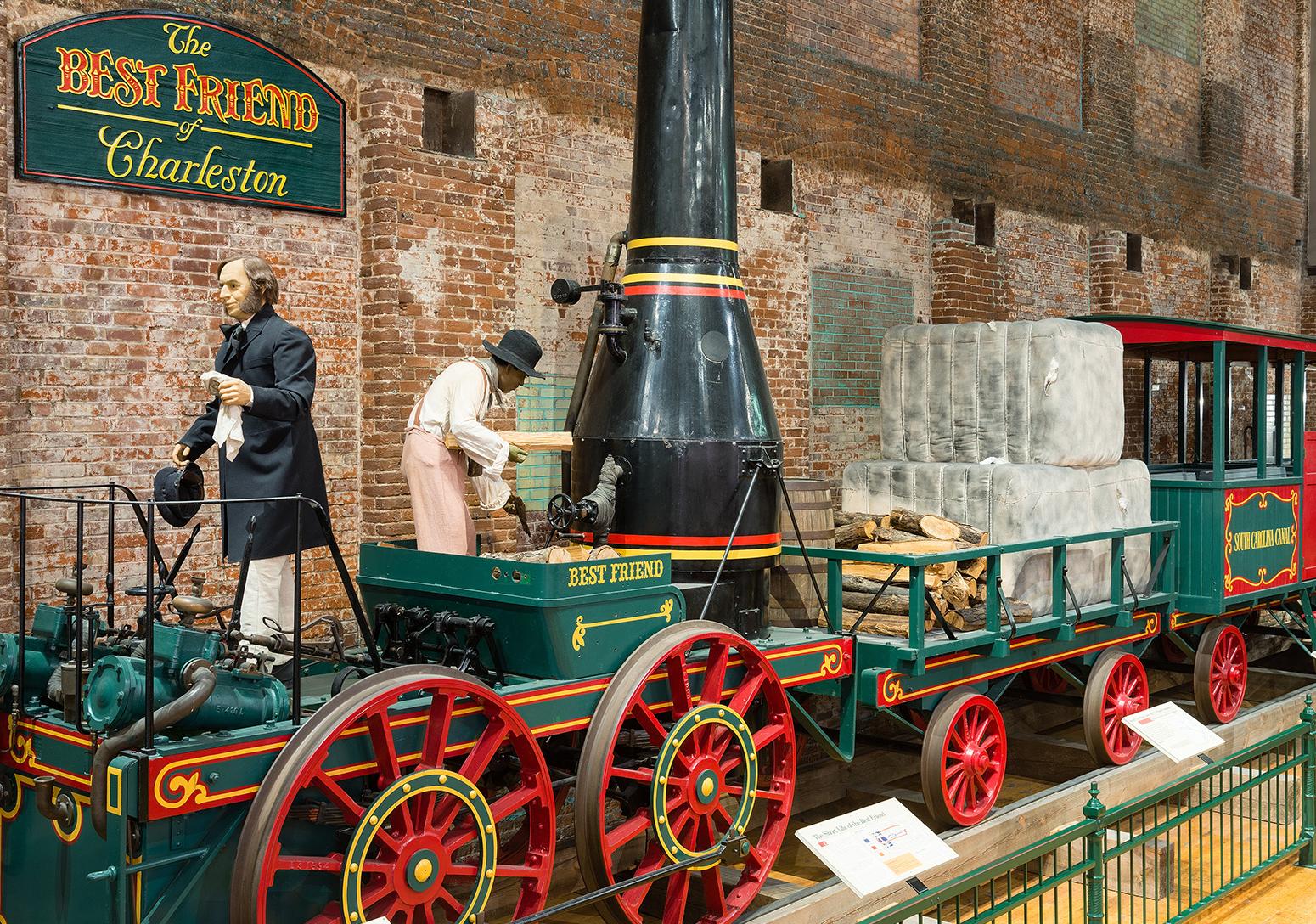 Full size train replica of The Best Friend of Charleston at the State Museum. Shows green train with red wheels and black smoke stack. 