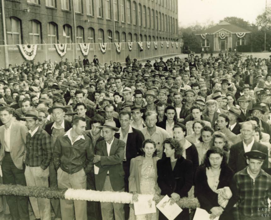 Large group gathered outside with side of textile mill building to the left with decorative patriotic banners on the windows