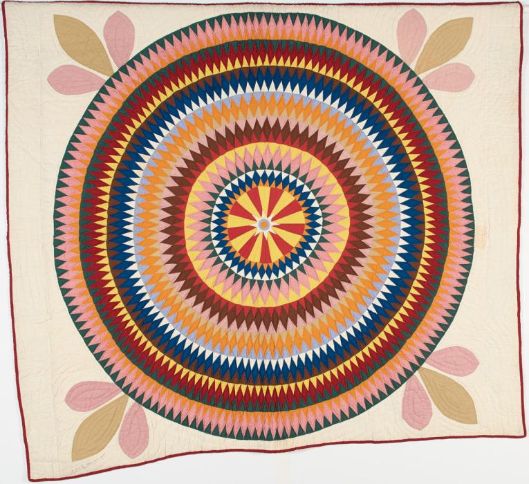 Quilt with cream backing and a light tan three leaf design at each corner. Center is made of concentric circles made of small diamond shaped fabric pieces in colors of blue, orange, red, brown, yellow and white.