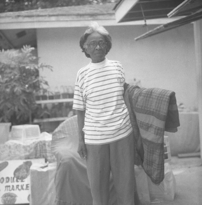 Black and white of woman standing on porch holding a quilt folded over her arm.