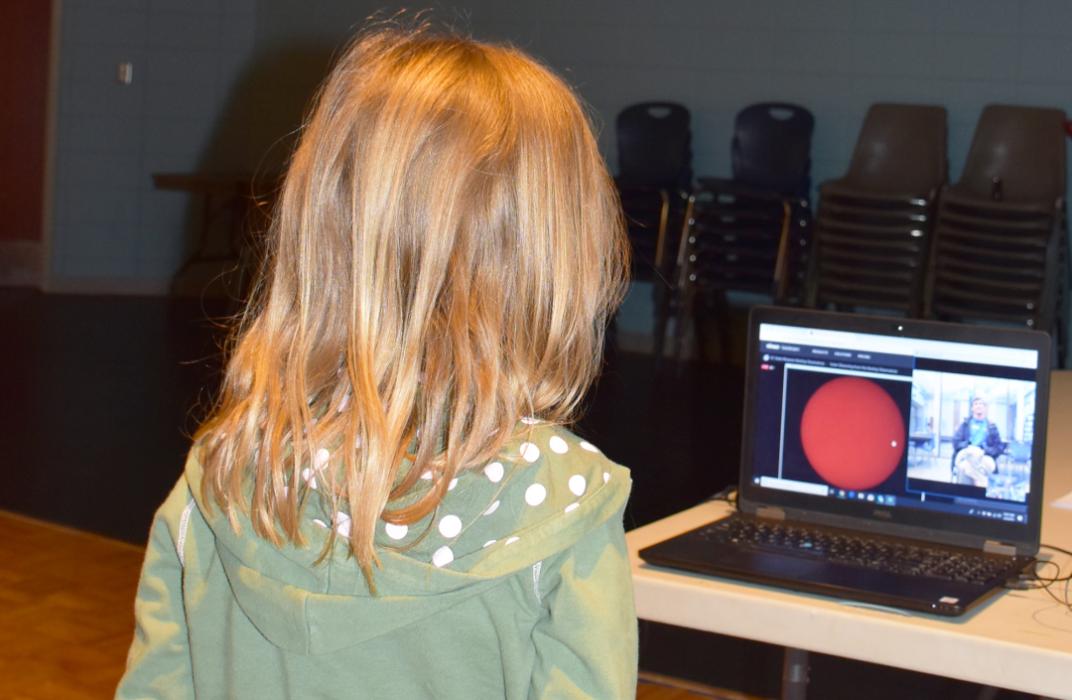 Young girl stands before laptop interacting live with an observatory educator on screen displaying an image of the Sun