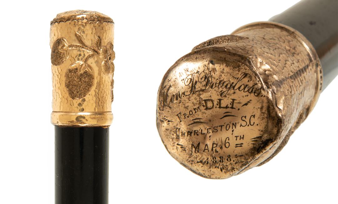 Showing the top of a walking stick. It is gold with fruit on the side and text on the top.