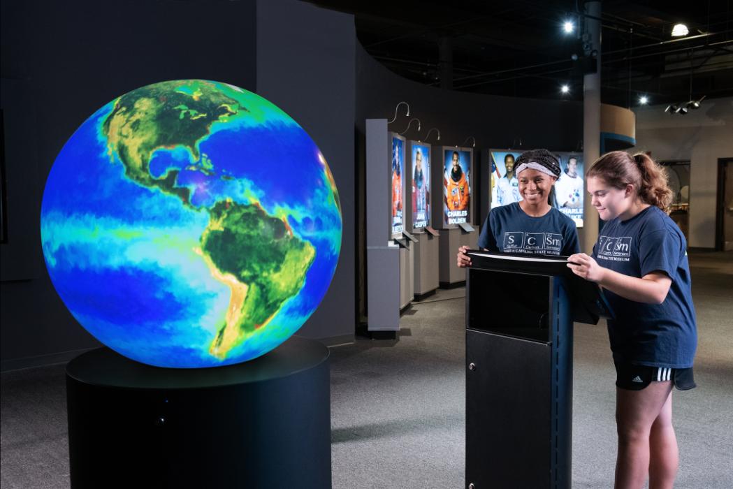 Two adolescent girls stand in front of a globe projecting an image of the Earth